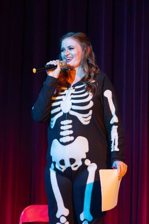 A performer in a skeleton costume sings on stage with dramatic lighting and dark backdrop.