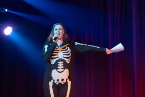 A performer in a skeleton costume singing on stage with dramatic blue lighting and red curtains in the background.