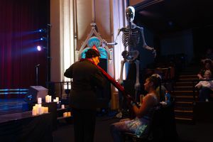 A theatrical scene with someone in a dark coat speaking with others near a skeleton prop and candles on stage.