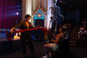 A musician performs with an electric guitar on a dark stage with Halloween decor including a glowing skeleton.