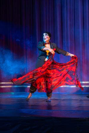 A performer in a black jacket twirls dramatically in a flowing red skirt on stage bathed in blue theatrical lighting.