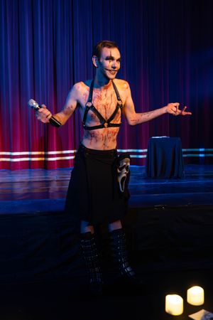 Performer in dark stage lighting with black costume and boots holds microphone during theatrical burlesque show.