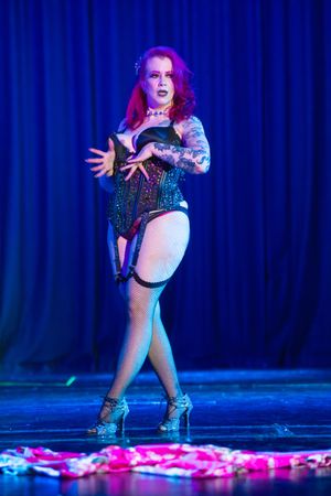 Burlesque performer with red hair in sparkly lingerie and heels performs on stage with blue lighting and curtains.