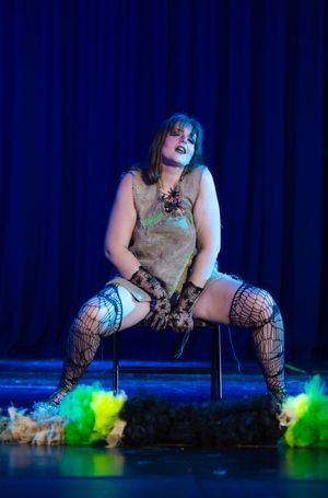 Cabaret dancer in fishnet stockings performs on chair with neon green props on dark stage.
