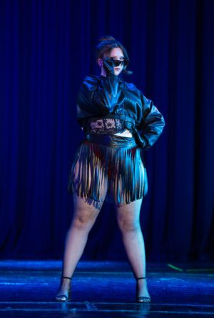 Performer in fringe outfit and sunglasses poses dramatically on dimly lit stage.
