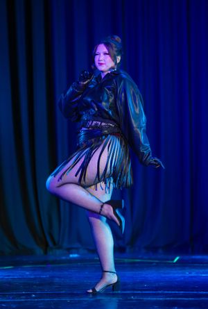 Dancer performs dramatic pose in dark costume against blue-lit curtain backdrop on stage.