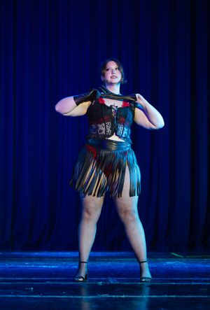 Performer in fringe costume and heels strikes dramatic pose on stage with blue curtain backdrop.