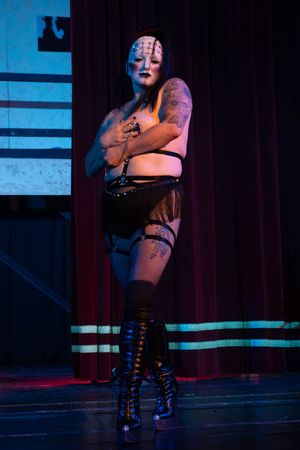 A burlesque performer in dark lighting poses dramatically on stage during a theatrical performance.