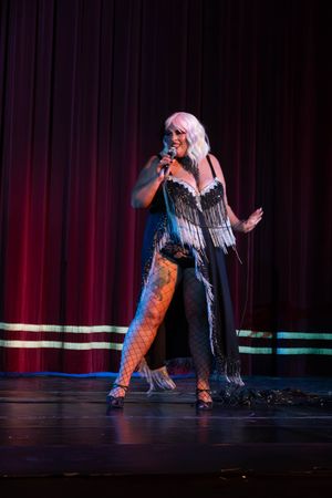 Performer on stage in a black fringe costume with fishnet stockings sings into microphone against red curtain backdrop.