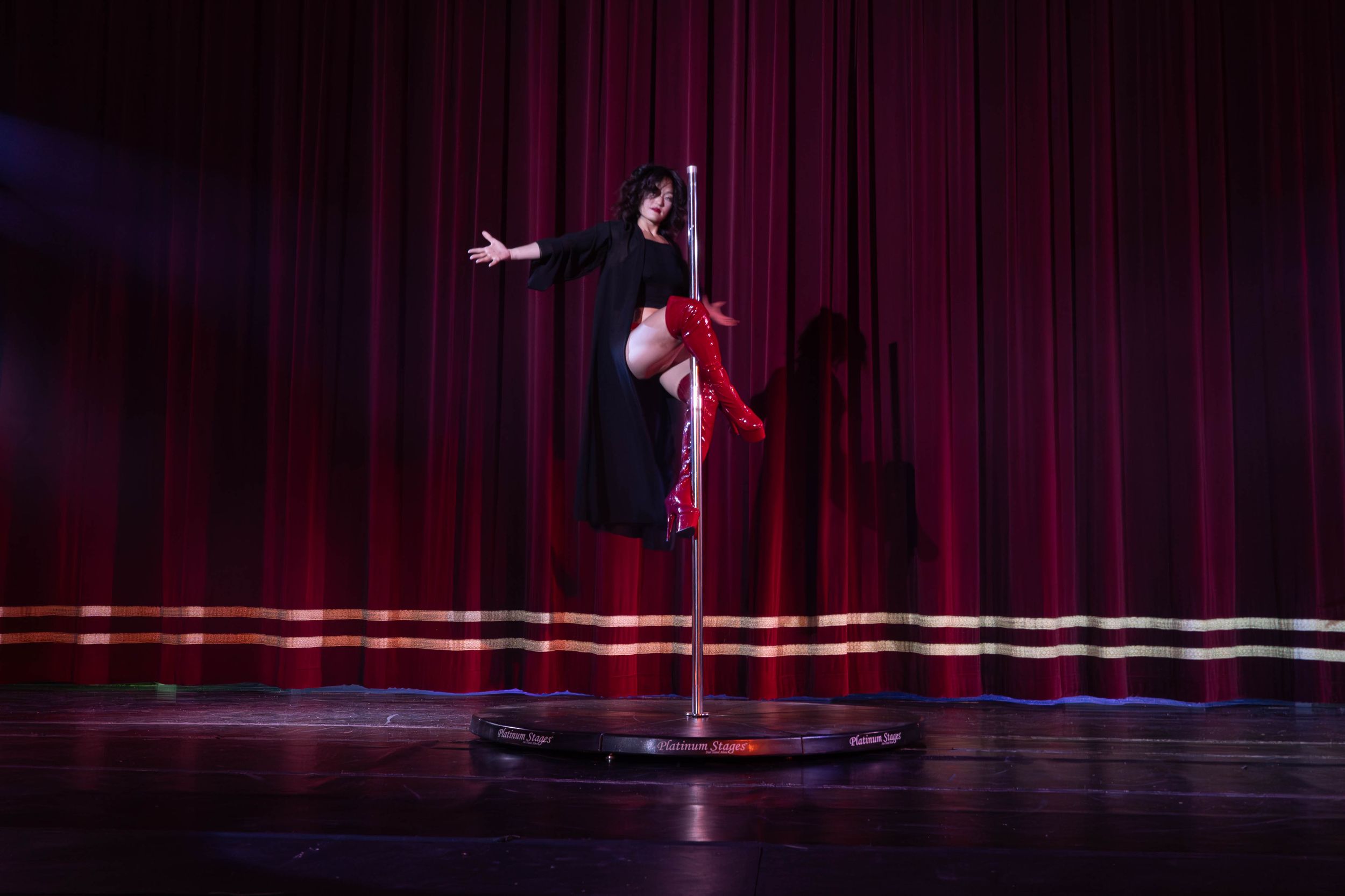 A performer in a black outfit does an acrobatic pole dance routine on stage with red curtains and dramatic lighting.