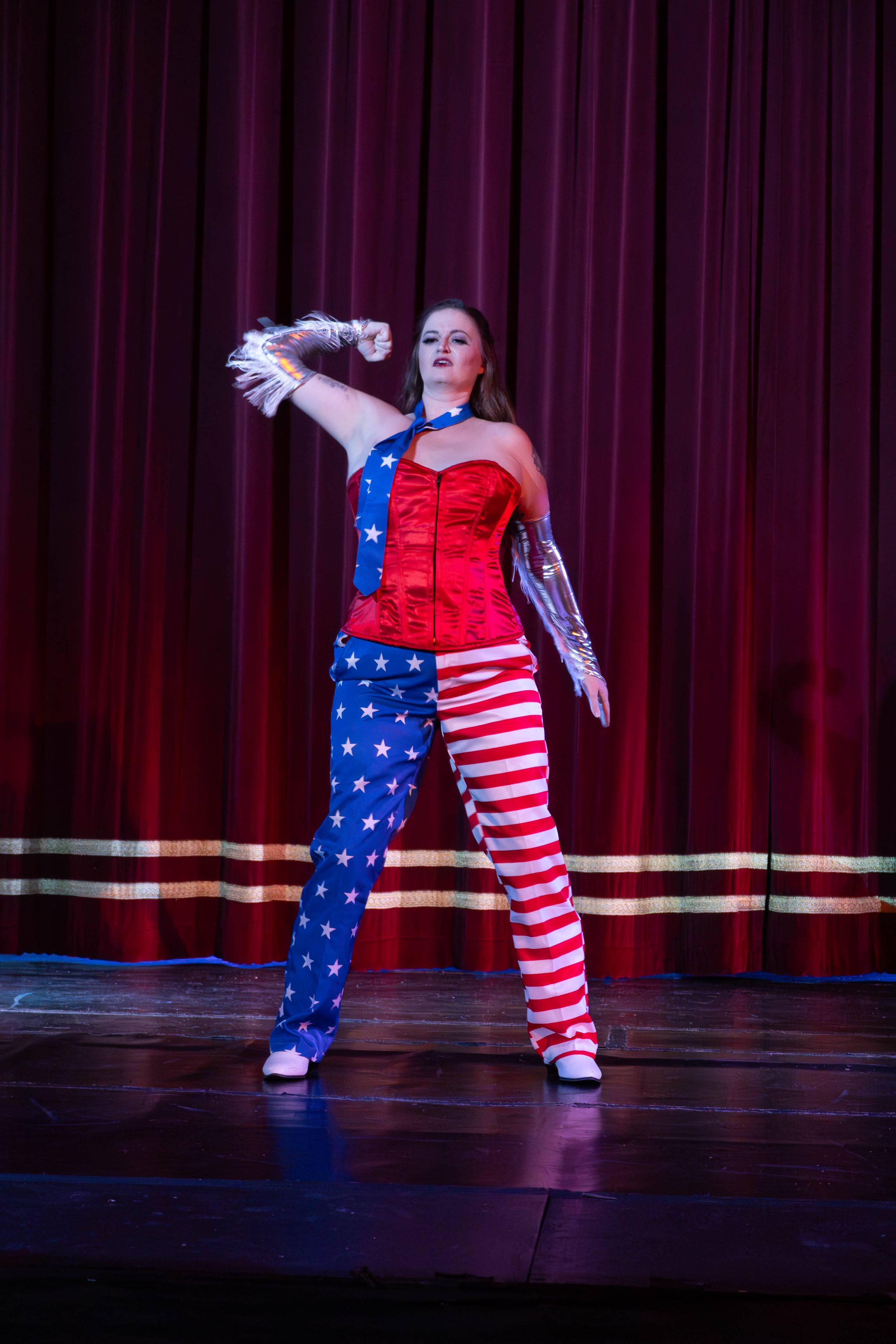 Performer in American flag-themed costume doing a dance move on dark stage with red curtain backdrop.