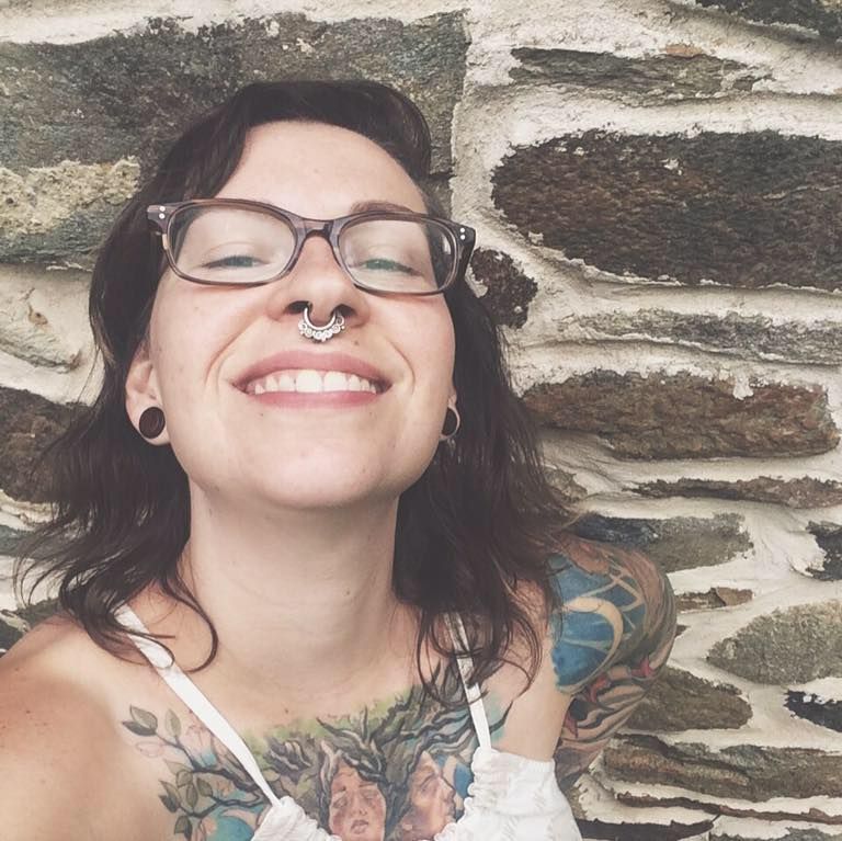 Playful selfie of Anne Kirk showing piercings and tattoos against a stone wall background.