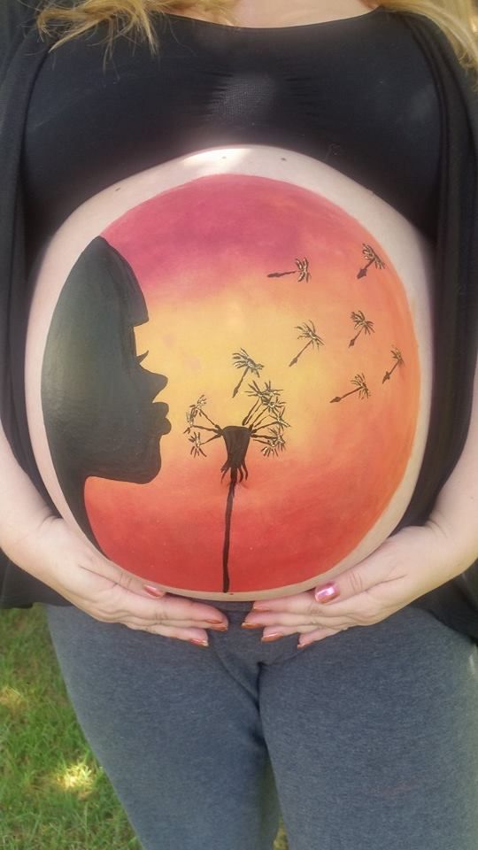 Silhouette of person blowing dandelion painted on pregnant belly against sunset background.
