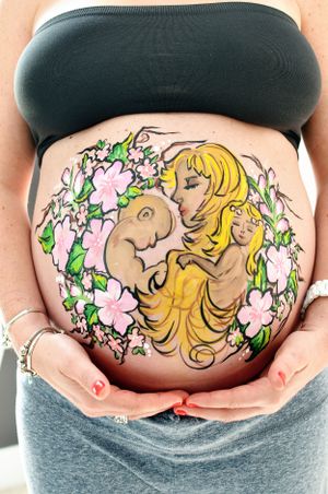 Artistic pregnancy belly painting featuring blonde figure surrounded by pink flowers.