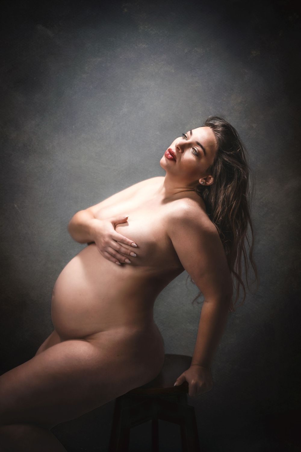 Artistic nude maternity portrait of a pregnant woman sitting gracefully on a stool against a dark studio background.