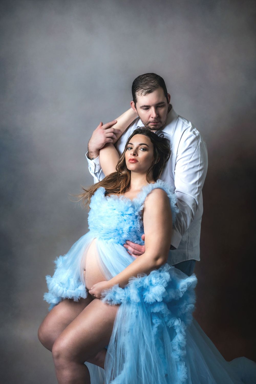 Pregnant woman in blue tulle gown poses with partner in white shirt during maternity photo shoot.