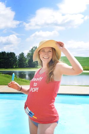 Pregnant woman in red tank top and sun hat poses by pool on sunny day.