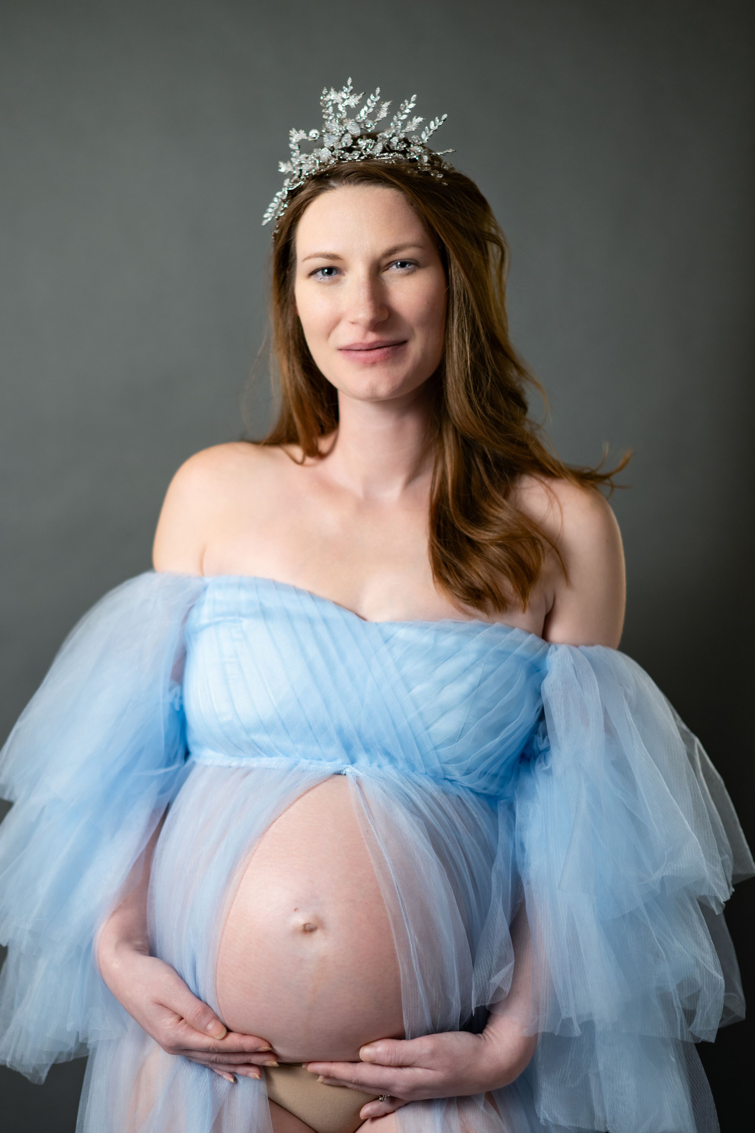 Maternity portrait in powder blue gown and crystal crown against grey background.