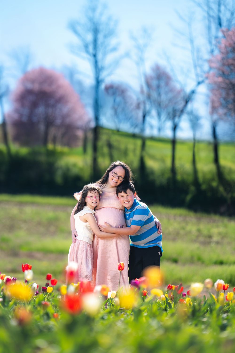 Pregnant woman in pink dress hugging two children in a colorful tulip field on a sunny spring day.