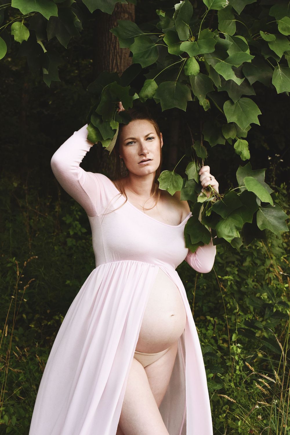 Maternity photo shoot in a white flowing dress against green foliage backdrop.