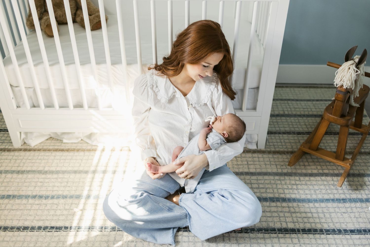 A mother with long red hair wearing light wide jeans holds her baby boy in the nursery while leaning on a white crib sitting on a rug.