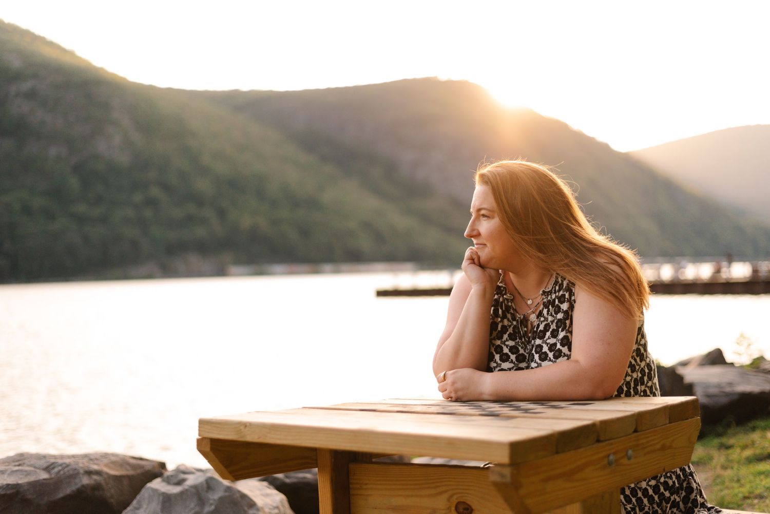 Person sits at wooden table by lakeside during golden hour sunset.
