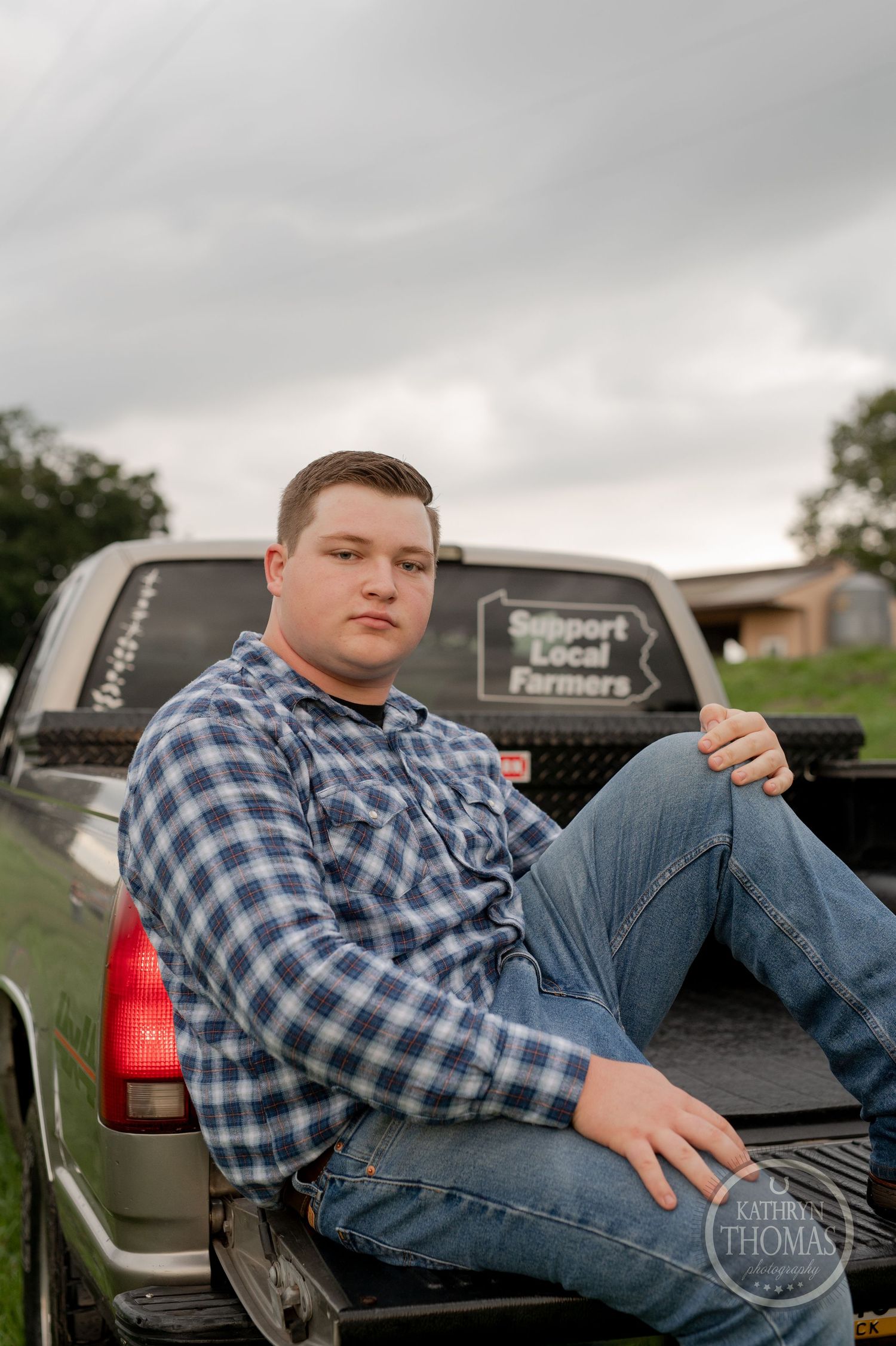 A person in plaid shirt sits in the bed of a pickup truck against cloudy skies.