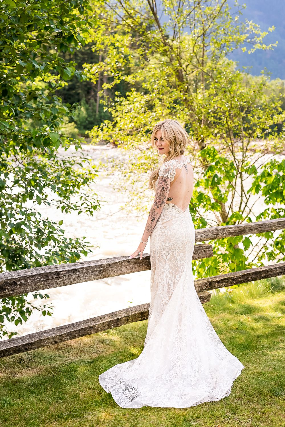 A bride shows off the back of her lacy wedding dress outside by the river.