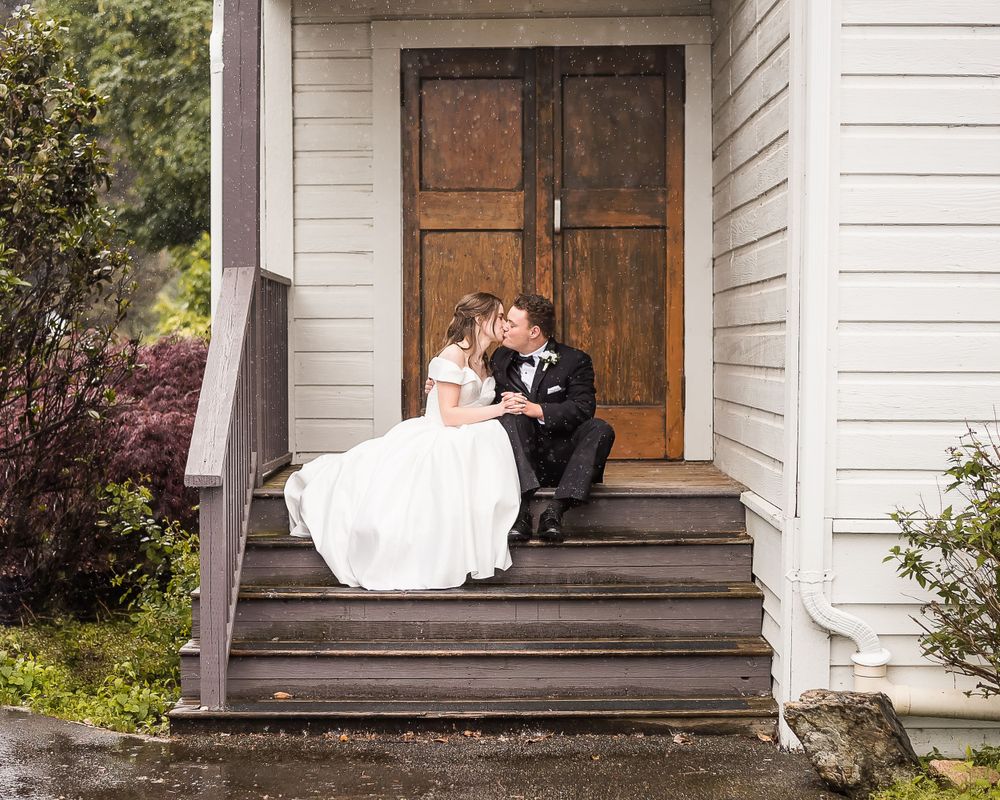 A bride and groom hide from the rain and kiss on the stairs of a church.