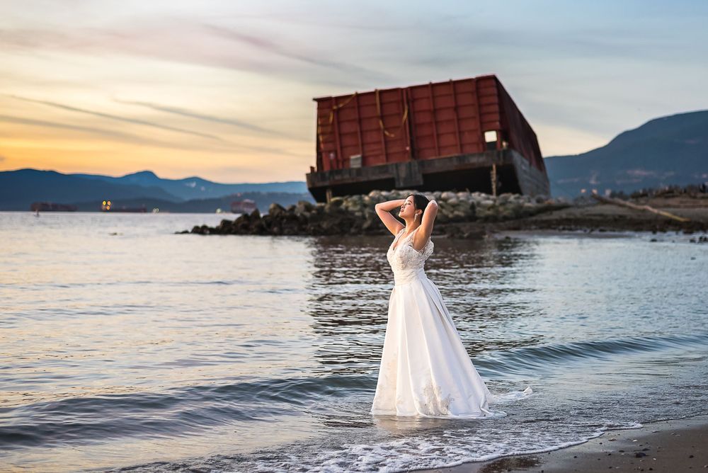 A bride in her wedding dress stands in the ocean at blue hour with a barge on the beach behind her.
