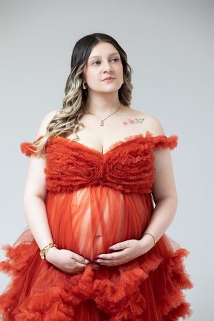 Pregnant woman in red ruffled tulle maternity gown cradling baby bump, floral shoulder tattoo, studio portrait.