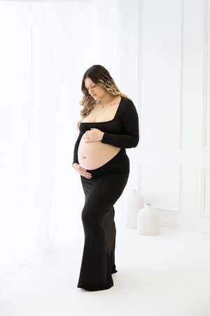 Pregnant woman in black crop top and skirt cradling baby bump in white studio maternity photo session.