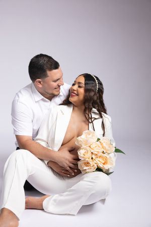 A couple in white attire shares an intimate moment while holding peach-colored roses against a light background.