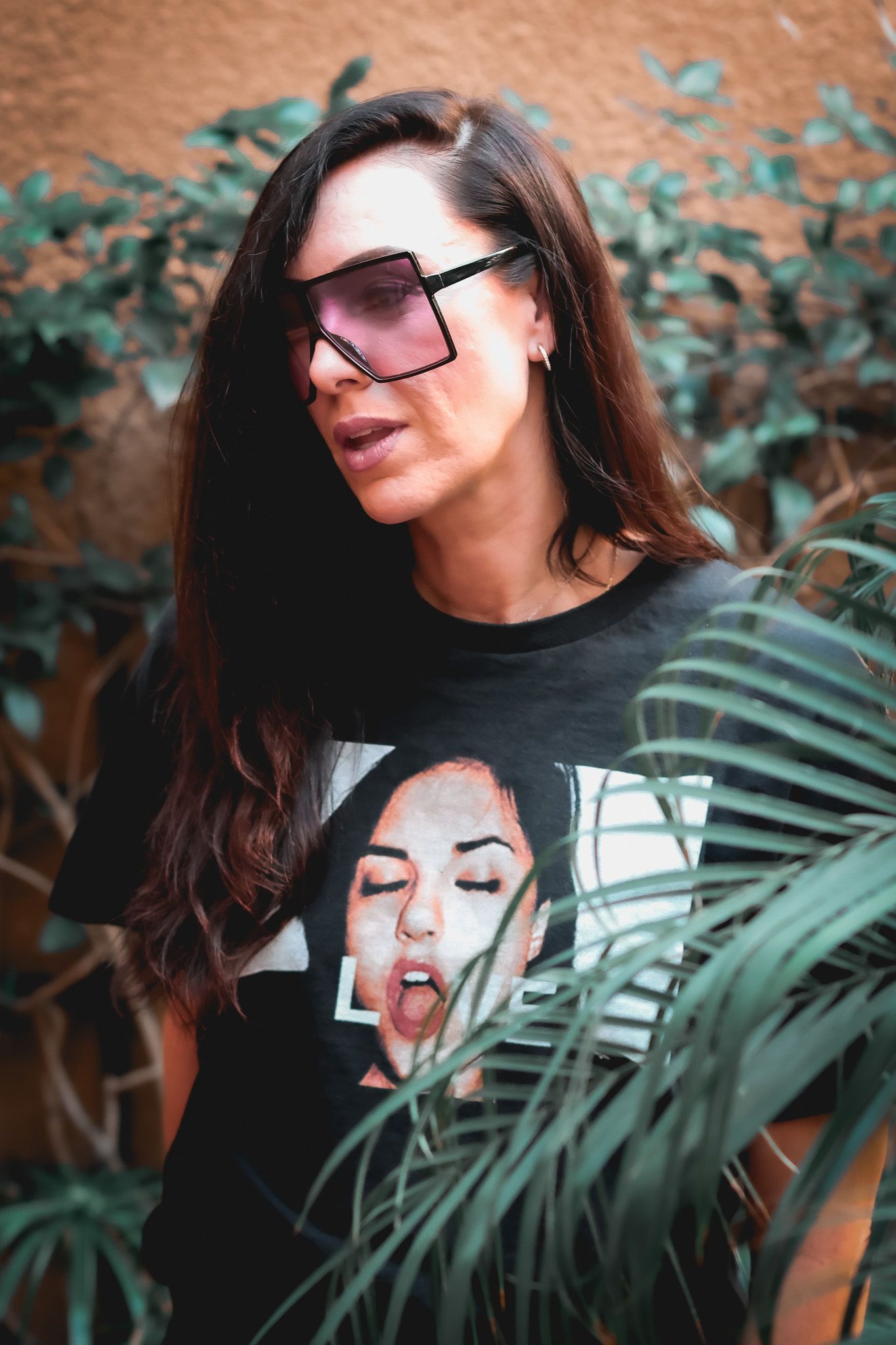 Fashion portrait of a woman wearing oversized pink sunglasses and a black graphic tee posing outdoors amid green plants.