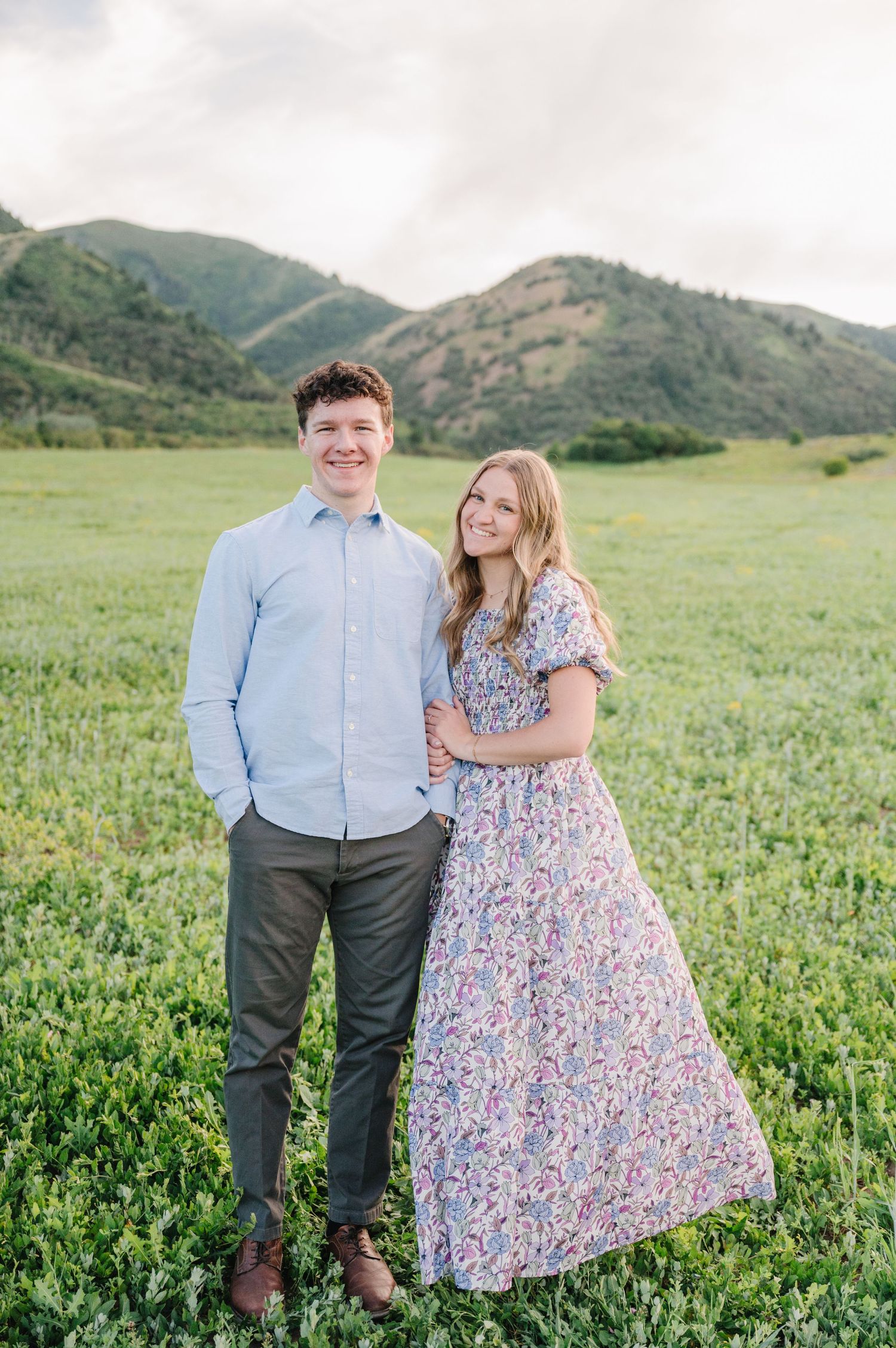Couple poses in a scenic field with rolling green mountains in the background during a spring photo session.