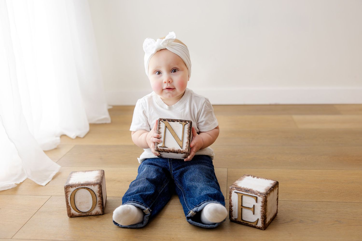 A baby wearing a white lace dress and pearl necklace reaches out while sitting against a white background.