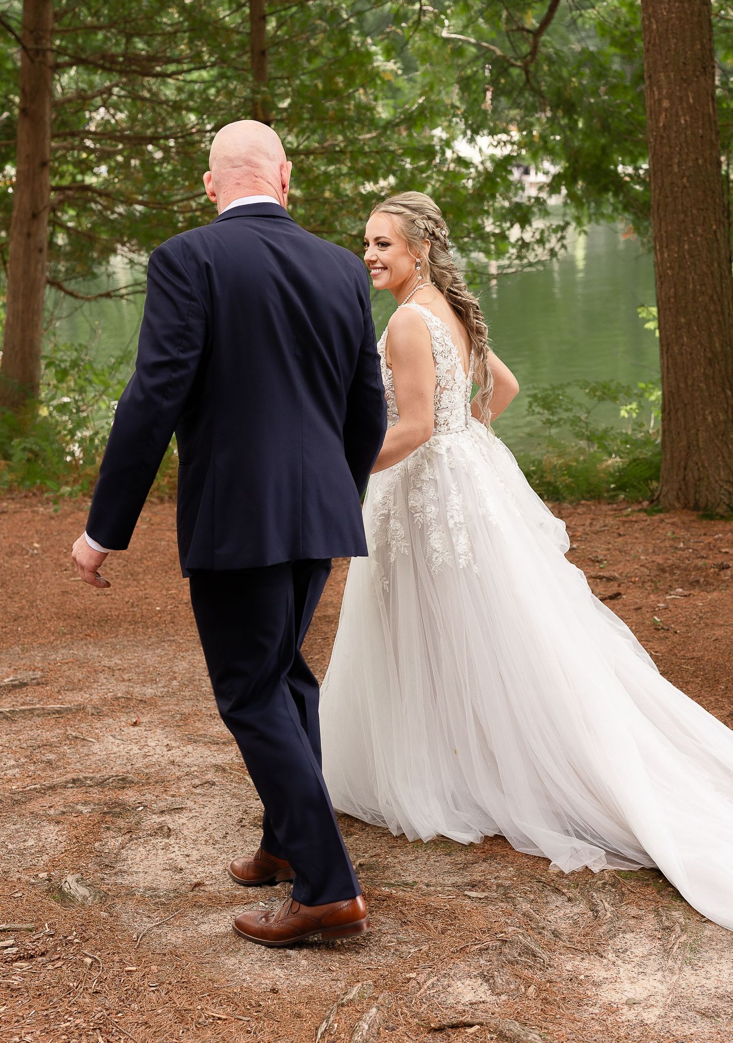 A bride and groom walk along a lakeside path surrounded by tall pine trees.