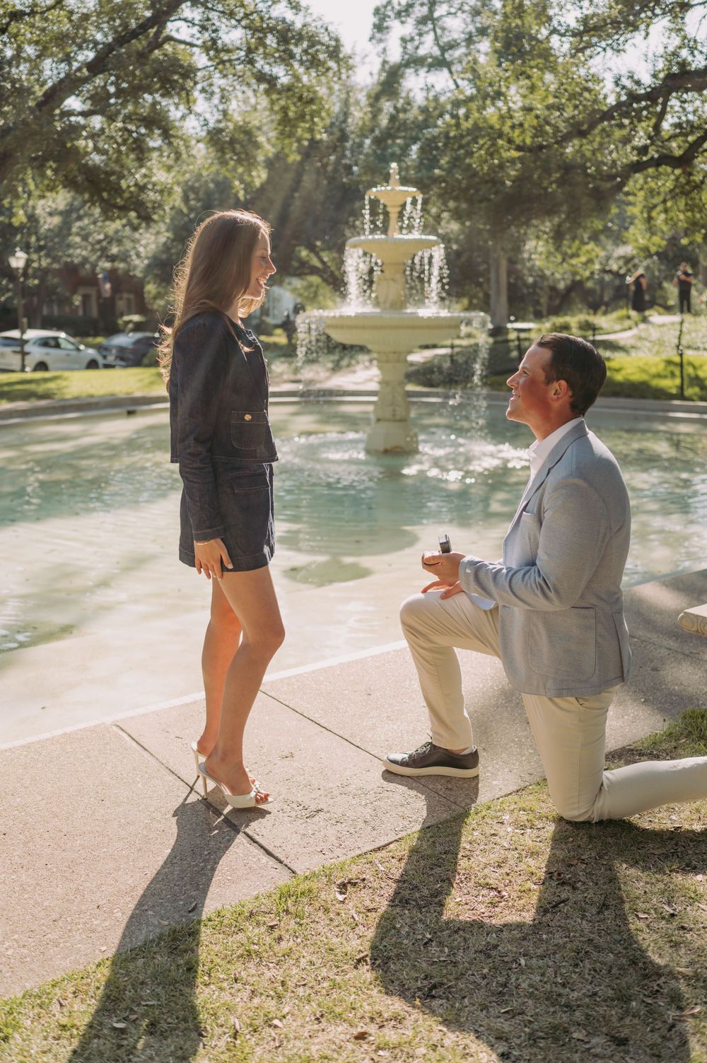 Romantic proposal scene by a garden fountain with sunlight streaming through the trees.