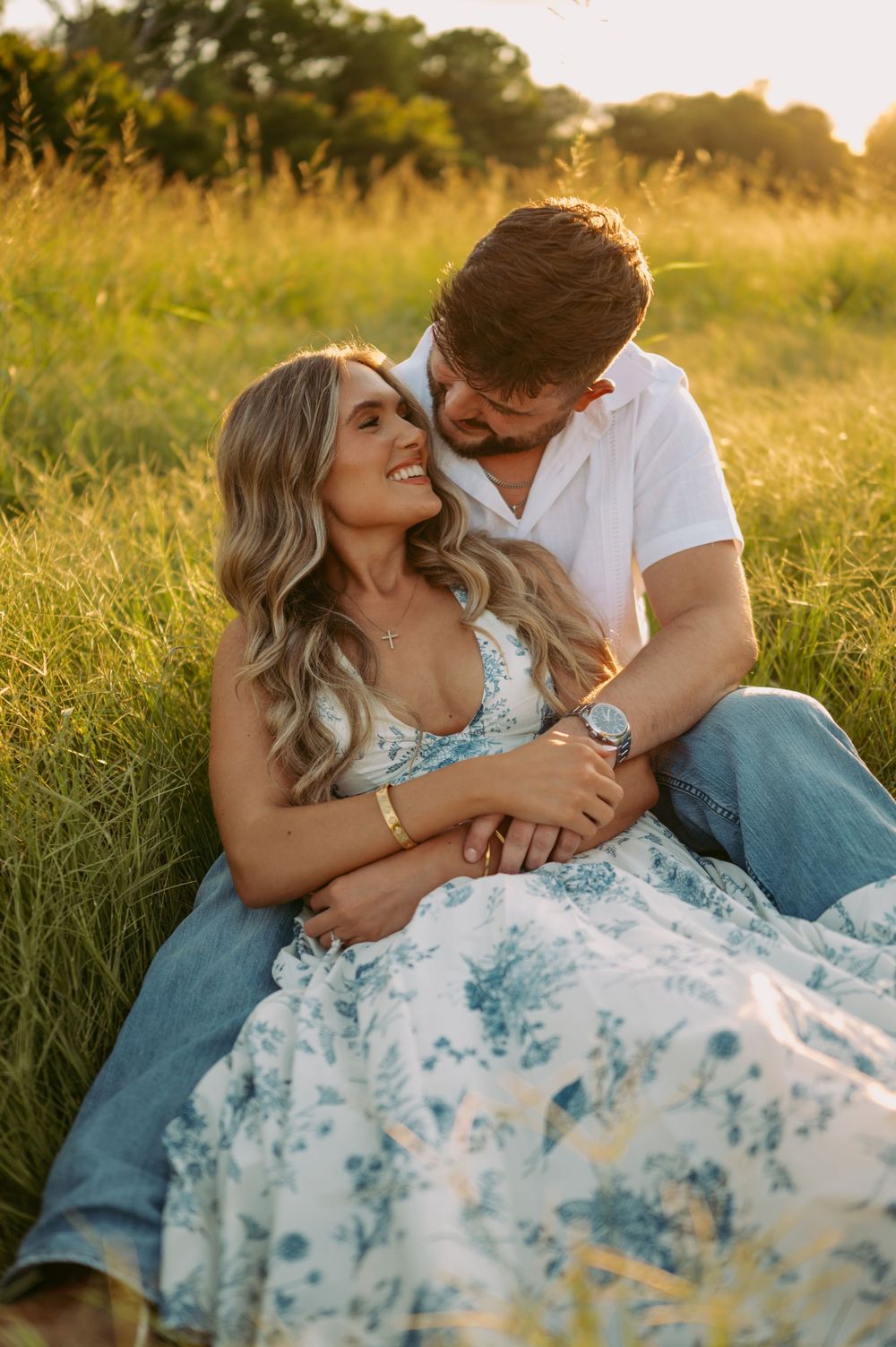 A couple shares intimate moments in a sunlit meadow wearing white and blue outfits during golden hour.