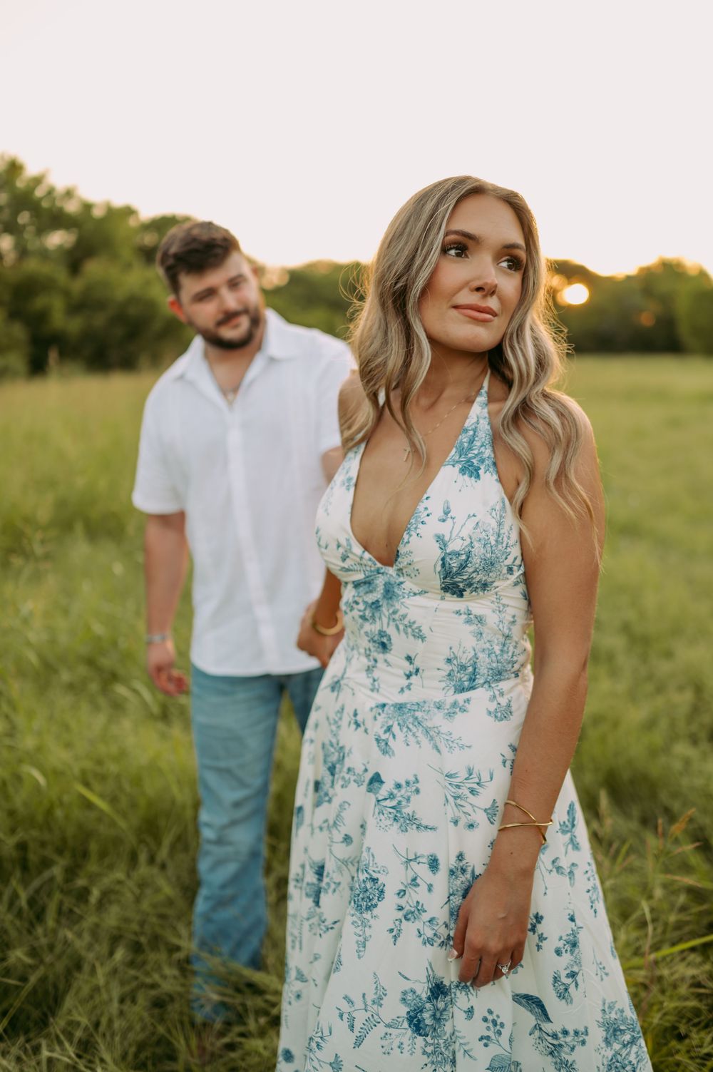 A romantic photoshoot in a grassy field features a flowing white and blue floral dress at sunset.