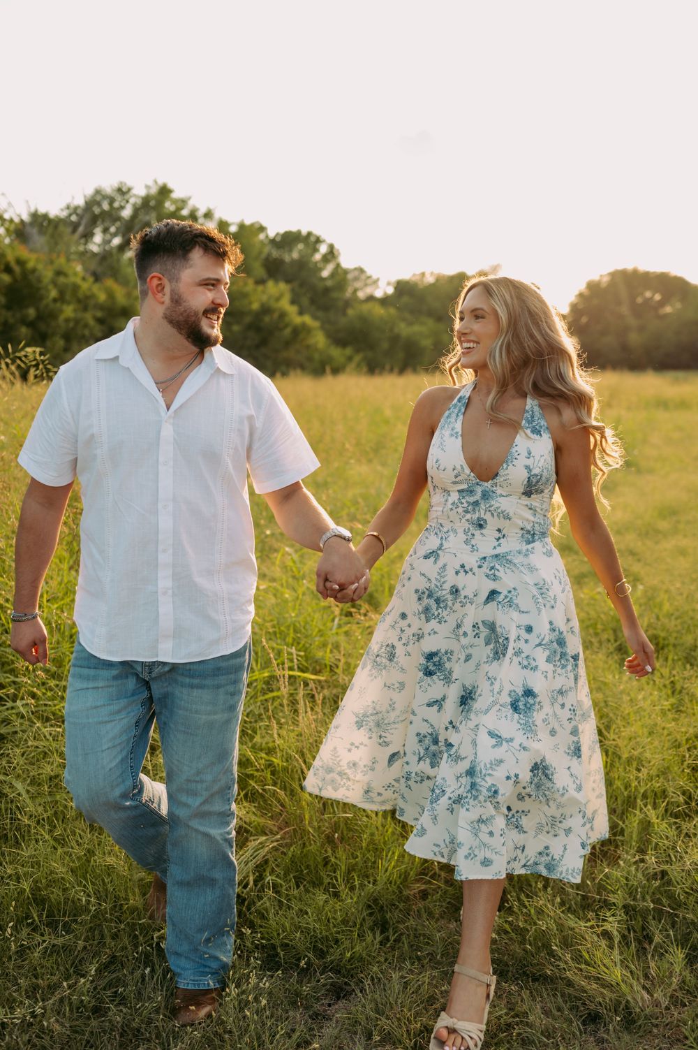 A couple holds hands while walking through a sunlit meadow at golden hour, she in a floral dress and he in casual attire.