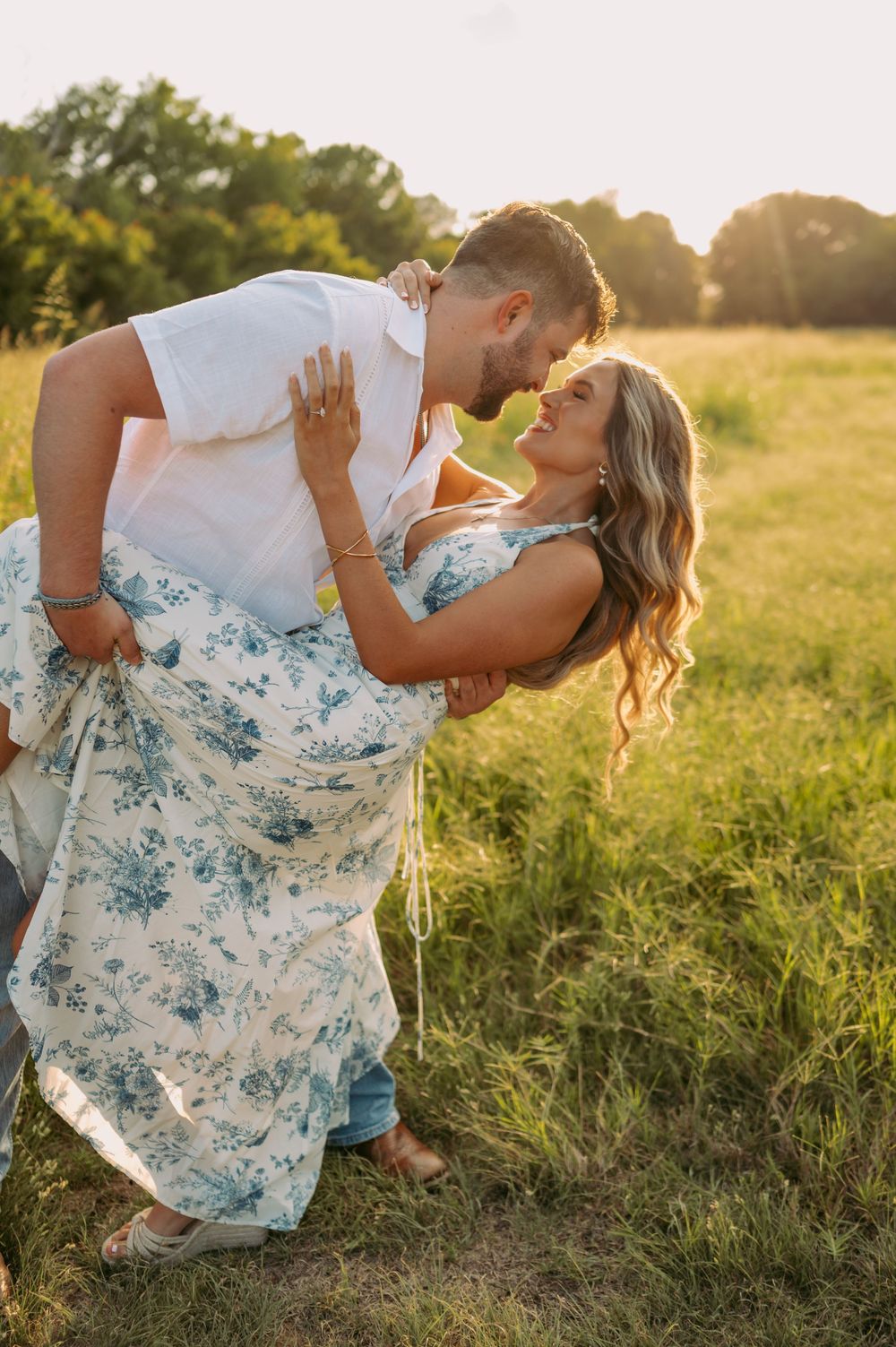 A couple dances together in a grassy field at sunset wearing coordinated white and blue floral outfits.