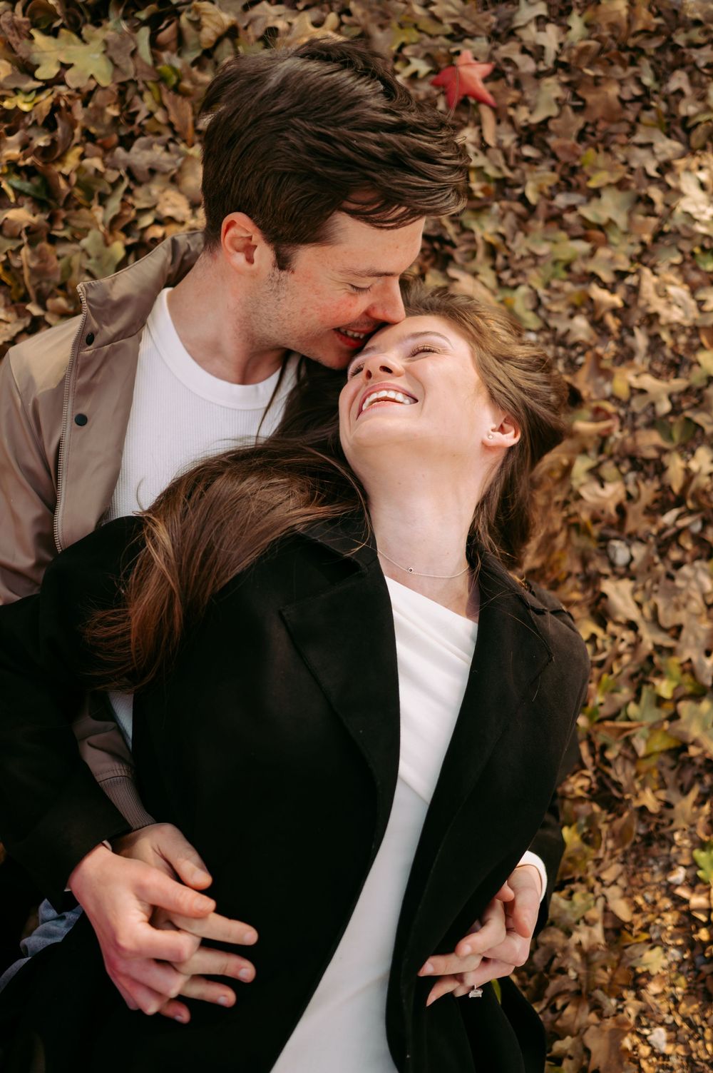 A couple shares an intimate moment lying on autumn leaves while wearing casual black and white clothing.