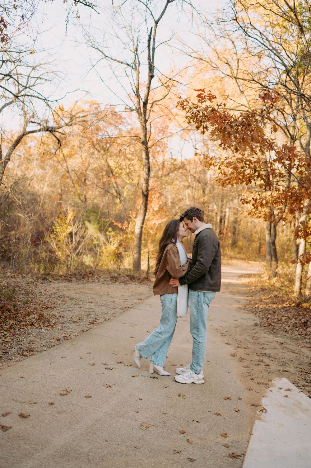 A couple embraces on a dirt path surrounded by autumn trees with golden foliage during sunset.