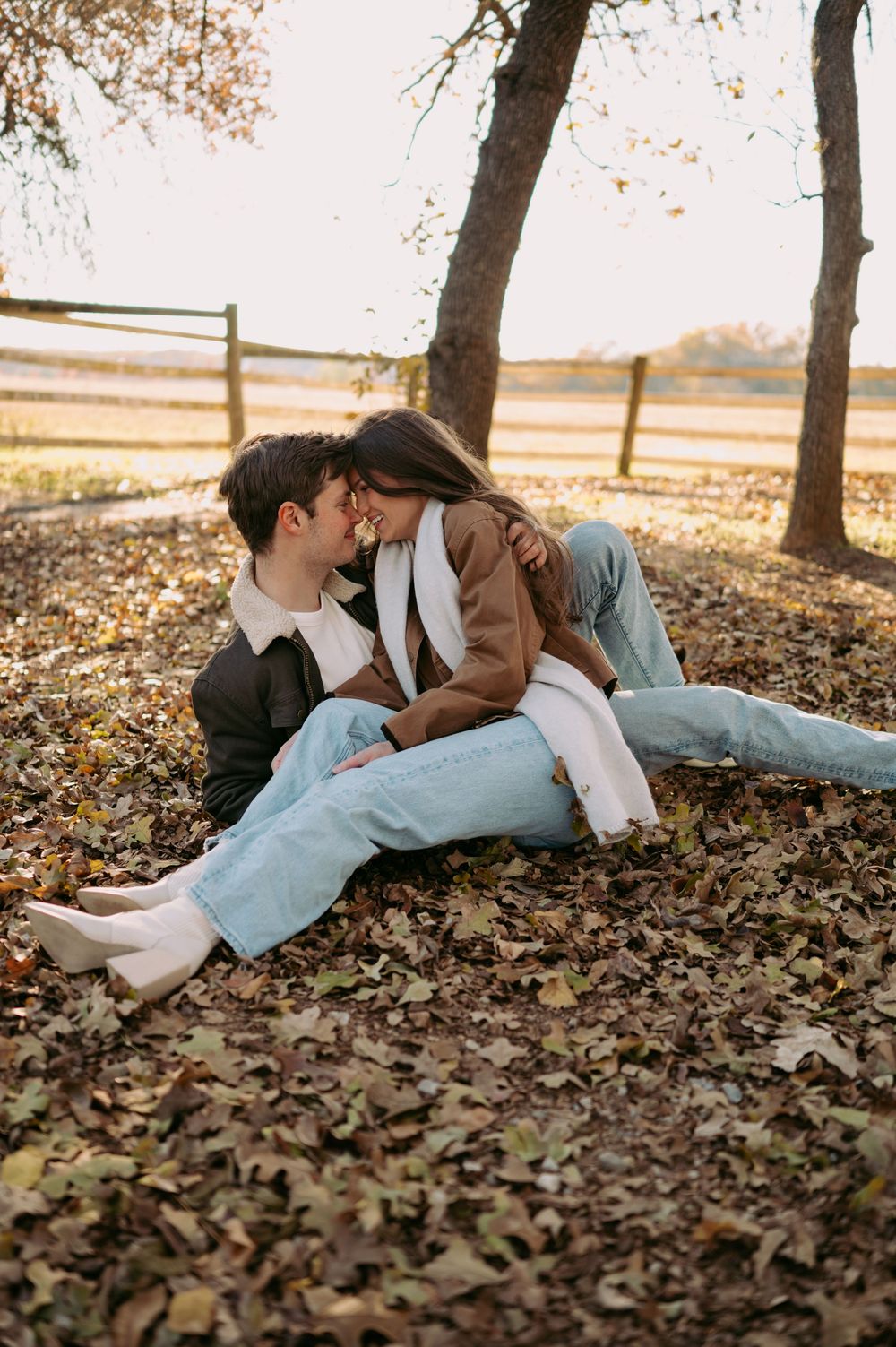 A couple lies together on fallen autumn leaves in a romantic outdoor setting during golden hour.