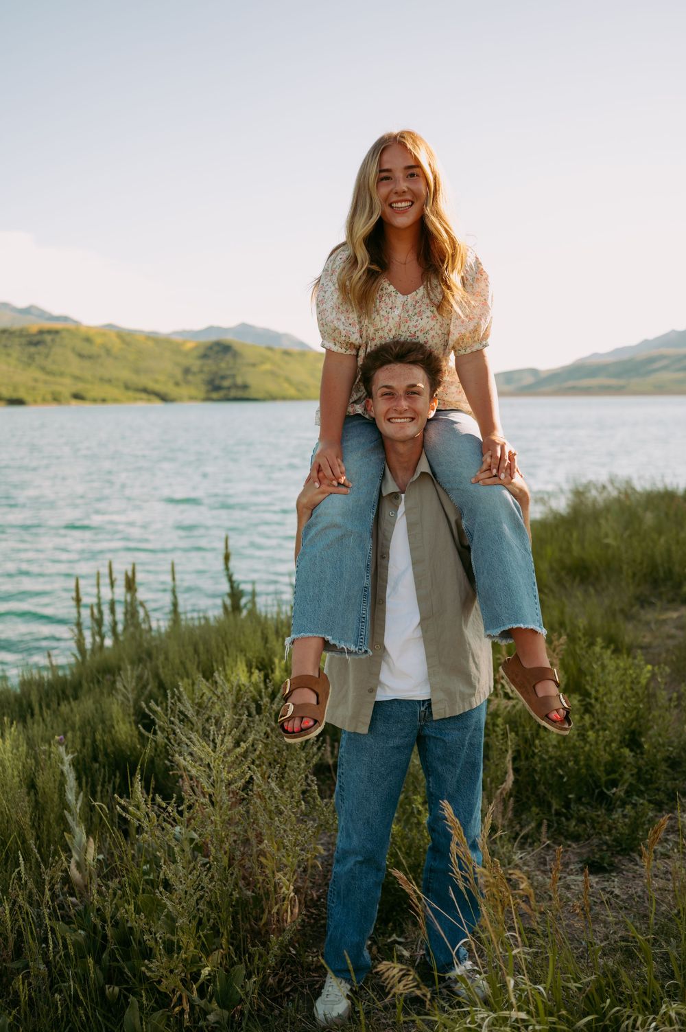 Two people stand together in a scenic mountain landscape with a lake and green vegetation at sunset.