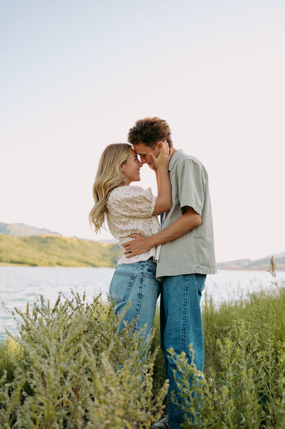 A couple embraces in tall grass by a lake during a romantic outdoor photoshoot.