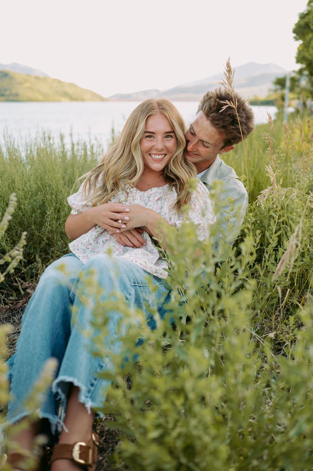Couple embraces in a field of tall grass and wildflowers near a mountain lake on a sunny afternoon.