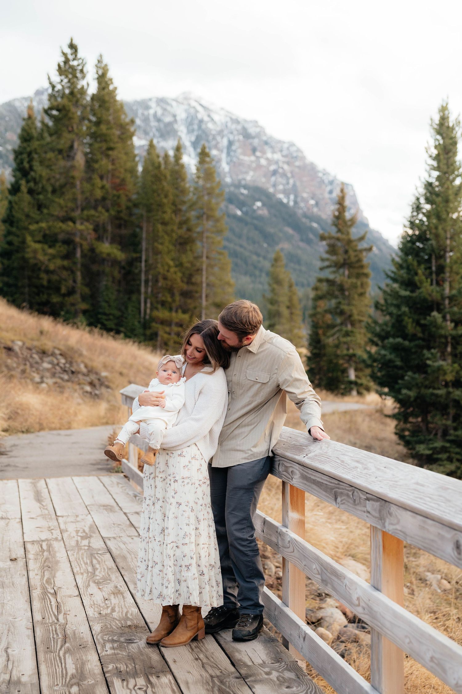 Young family with infant on rustic wooden bridge surrounded by pine trees and snow-dusted mountain.