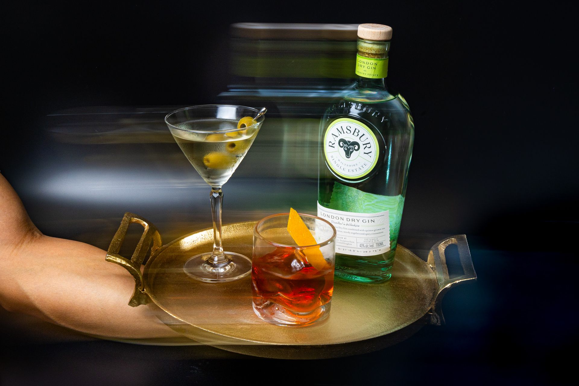 A martini and negroni cocktail on a gold tray beside a green gin bottle, elegantly served by a bartender.